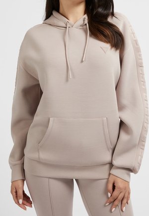Kobieta w beżowej bluzie z kapturem z troczkami i przednią kieszenią, w zestawie z dopasowanymi legginsami, stoi na jednolitym tle.