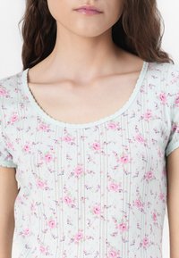 Top azzurro chiaro a manica corta con motivo floreale in rosa e verde. Tessuto strutturato con bordi ondulati sul collo e sulle maniche.