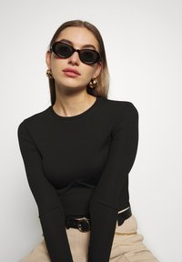 Top negro de manga larga acanalada combinado con un cinturón y pantalones beige; lleva gafas de sol ovaladas de carey y pendientes de aro dorados.