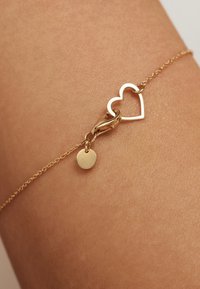 Gouden armband met een hartvormige bedel en een ronde tag. De ketting is Delicaat met in elkaar grijpende schakels die op de huid rusten.