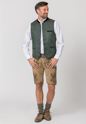 Mann trägt traditionelle bayerische Tracht mit grünem Weste, weißem Hemd, bestickter Lederhose, grauen Socken und braunen Schuhen, steht und lächelt.