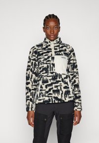 Pullover in pile con stampa crema e blu navy, dotato di collo a zip, tasca sul petto bianca e tessuto strutturato. Sotto sono indossati pantaloni cargo neri.