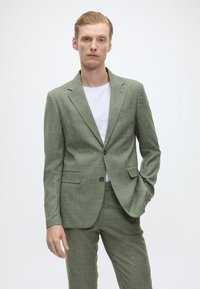Helles grünes Anzug aus strukturiertem Stoff, mit einem einzelnen Knopfverschluss, Reverskragen und zwei vorderen Taschen, kombiniert mit einem weißen Hemd.