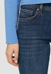 Jasnoniebieska koszula z długim rękawem w połączeniu z ciemnoniebieskimi dżinsami. Dżinsy mają klasyczny pięciokieszeniowy fason, z pomarańczowymi przeszyciami i teksturowanym materiałem.