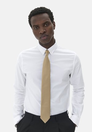 Chemise blanche à manches longues, associée à une cravate beige unie. Le modèle porte un pantalon sombre et se tient devant un fond neutre.