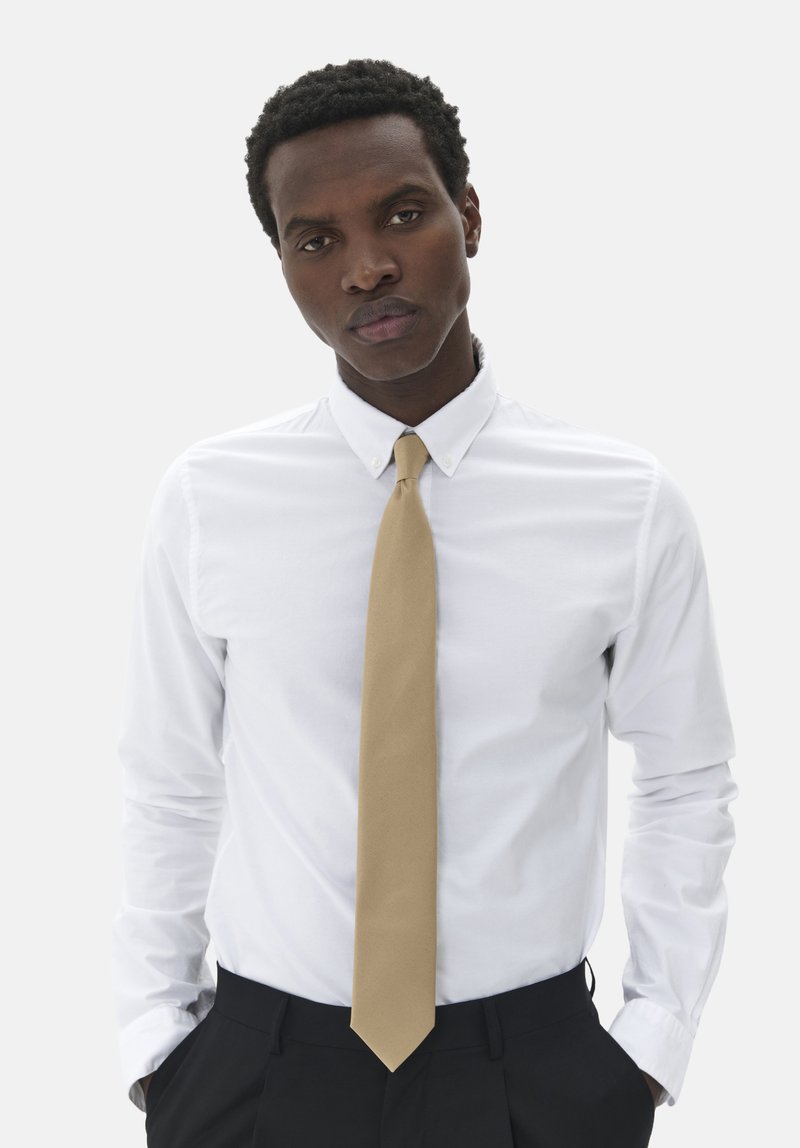 Vit skjorta med lång ärm, i kombination med ett enfärgat beige slips. Modellen har på sig mörka byxor och står mot en neutral bakgrund.