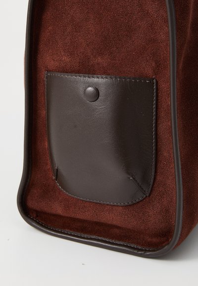 Sac en suede marron avec une poche en cuir foncé dotée d'une fermeture à pression, encadrée de passepoil marron. La surface est douce avec une finition texturée.