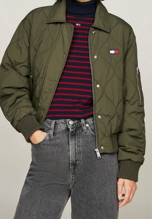 Persoon draagt een olijfgroene doorgestikte bomberjas over een marineblauwe en rood gestreepte coltrui, gecombineerd met grijze jeans met hoge taille.