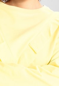 Top in cotone giallo con una vestibilità rilassata, caratterizzato da un collo a V e due tasche frontali. Texture morbida e design minimalista.