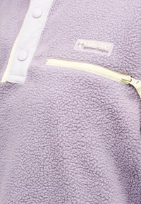 Ελαφρώς μωβ φλις ένδυμα με υφή, με ανοικτού κίτρινου χρώματος τσέπη με φερμουάρ και λωρίδα με κουμπιά, στολισμένο με το λογότυπο της Columbia Sportswear Company.