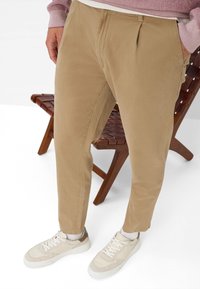 Beige op maat gemaakte broek met voorplooien, van katoen, met een taps toelopende pasvorm, gecombineerd met lichtgekleurde sneakers. Houten stoel op de achtergrond.