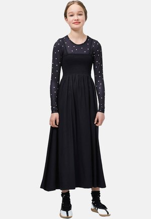 Young girl standing, wearing black long dress layered over a sheer black top with white heart patterns, and black and white sneakers.