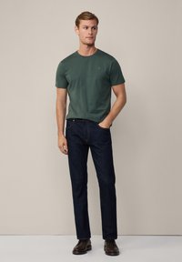 Tmavozelené tričko s krátkym rukávom, slim fit, spojené s tmavomodrými džínsami s rovnými nohavicami a hnedými koženými čižmami na neutrálnom pozadí.
