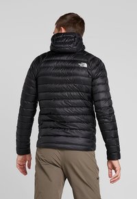 Czarna kurtka puchowa z wysokim kołnierzem, poziomym pikowaniem i logo na górnej części pleców. Gładka, błyszcząca tkanina.