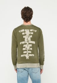 Vīrietis ar cirtainiem brūniem matiem, ģērbies olīvzaļā džemperī ar baltu abstraktu tekstu uz muguras, valkājot gaiši zilas džinsas, stāv pret baltu fonu.