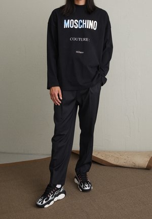 Person iført sort Moschino langærmet skjorte, sorte bukser og sorte og hvide sneakers med flammemønster, stående indendørs.