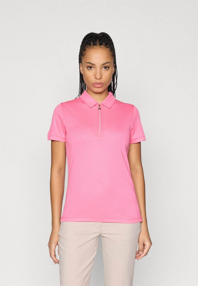 Polo majica v svetlo roza barvi, kratki rokavi, z zadrgo pri ovratniku. Narejena iz teksturirane tkanine. V kombinaciji z svetlo bež hlačami.