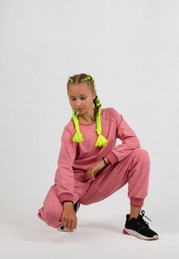 Rosa Sportanzug mit gerippten Bündchen, lockerer Passform und strukturiertem Stoff. Neon-grüne Zöpfe mit Pompon-Enden. Schwarze Sneakers.