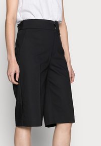 Žena v černých kraťasech s vysokým pasem, sahajících ke kolenům, které mají boční kapsy, a s bílé texturované bezrukávové blůze zasunuté v pase.