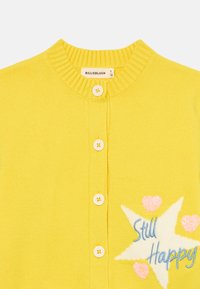 Gul stickad cardigan med rund hals, fem vita knappar och en stjärndesign som har "Still Happy" i blå och rosa accenter.