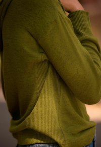 Oliven grøn sweater med en blød tekstur, der har ribbede detaljer på siderne og et asymmetrisk design. Nærbillede af tøjet.