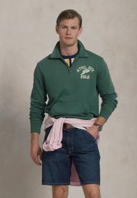 Felpa verde con zip e grafica "ATHL.DIV. POLO", abbinata a pantaloni di jeans e una maglietta a righe rosa legata in vita.