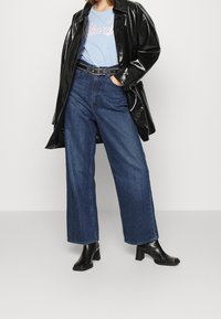 Mujer vestida con jeans de pierna ancha de color azul, botas de tacón negras, camiseta gráfica de color azul claro, cinturón negro y chaqueta de cuero negro brillante, de pie con una mano en el bolsillo.