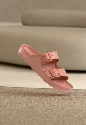 Sandale slide rose unique avec deux boucles réglables, semelle texturée, flottant au-dessus d'un tapis beige devant un canapé neutre.