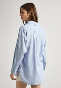 Camicia azzurra chiara con maniche lunghe e yoke posteriore. Presenta un orlo curvo e una piccola etichetta vicino al colletto. Tessuto morbido.