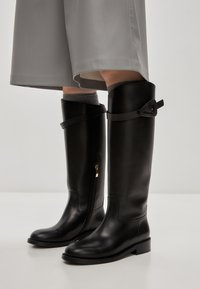 Cizme de călărie negre, până la genunchi, din piele netedă, cu fermoar pe lateral și detalii decorative cu curea în partea de sus. Talpă plată cu un călcâi ușor.