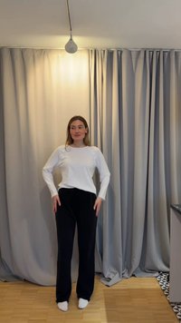 Kvinne står barbeint på tregulv foran lys og mørkegrå gardiner, iført hvit langermet skjorte og svarte bukser.