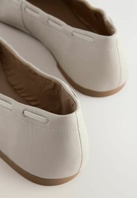 Baleto balerinos iš balto odos su lygiu paviršiumi, apvalios nosies dizainu ir kontrastinga rudos gumos padu. Dekoratyviniai šoniniai įpjovimai detalėms.