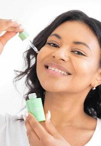 Grön glasdroppflaska märkt "Retinol & Vitamin C", med en klar droppare; en person som håller den, ler med strålande hud.