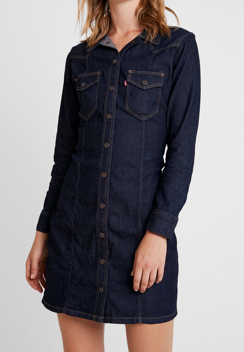 Femme portant une robe en jean bleu foncé boutonnée, manches longues et poches à rabat sur la poitrine, debout devant un fond uni.