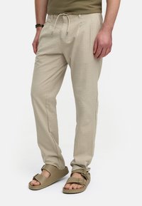 Beige texturerade byxor med dragsko i midjan; två sidofickor; kombinerade med bruna sandaler. Tyget har ett subtilt mönster.