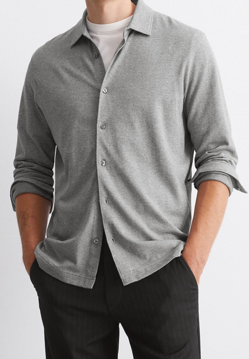 Homme portant une chemise grise boutonnée avec les manches retroussées et les mains dans les poches, associée à un pantalon noir à fines rayures.