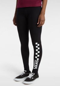 Leggings neri con motivo a scacchi bianchi e branding sulla gamba sinistra, indossati con scarpe nere con suole e lacci bianchi.
