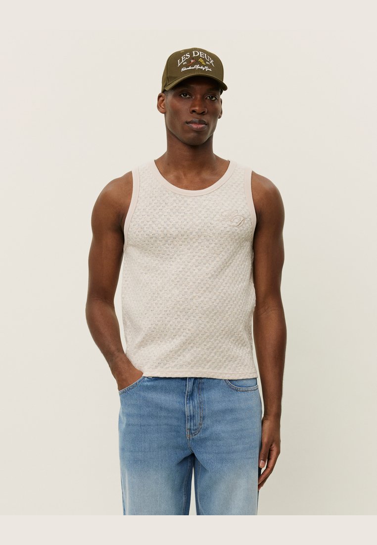 Ljusbeige linne med texturerad tyg, rund halsringning och ribbade kanter, kombinerat med blå denimjeans. Avslappnad stil med minimalistiska detaljer.