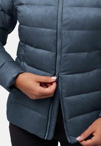 Die Hände einer Person schließen eine dunkelblaue, gesteppte Steppjacke über eine schwarze Hose vor einem schlichten Hintergrund.