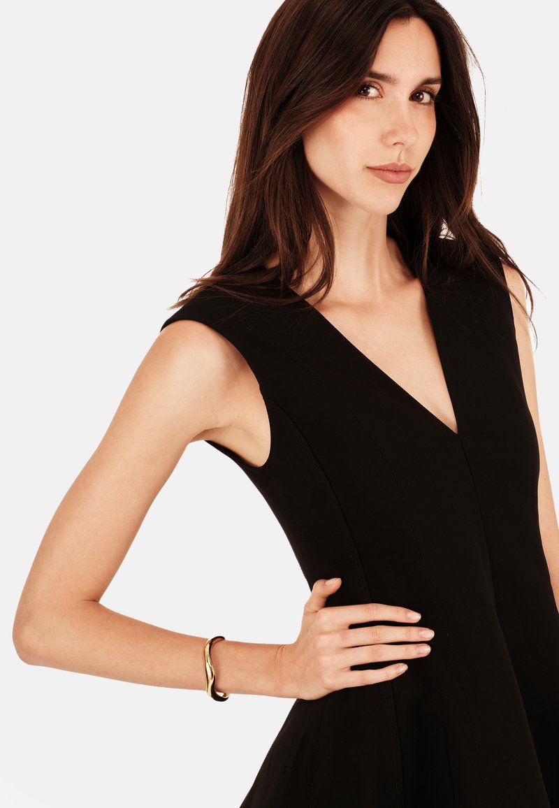 Vestido negro sin mangas con diseño de escote en V y cuerpo ajustado. Modelo lleva una pulsera dorada en la muñeca. Textura suave, silueta elegante.
