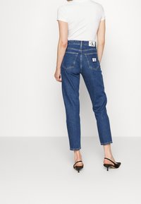 Korkeavyötäröiset, suorat siniset farkut, joissa on kaksi takataskua ja logoplässi vyötäröllä, yhdistettynä valkoiseen, vartalonmyötäiseen toppiin.