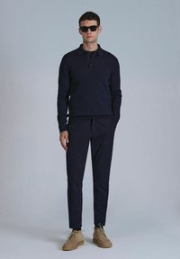 Marineblaues, geripptes Langarm-Polo und passende, maßgeschneiderte Hose, kombiniert mit beigen Wildlederschuhen. Das Modell trägt schwarze Sonnenbrillen vor einem neutralen Hintergrund.