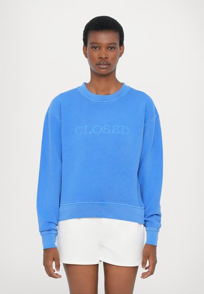 Sweatshirt bleu avec "CLOSED" brodé en fil léger, poignets et ourlet côtelés, coupe ample, porté sur un short blanc. Texture en coton lisse.