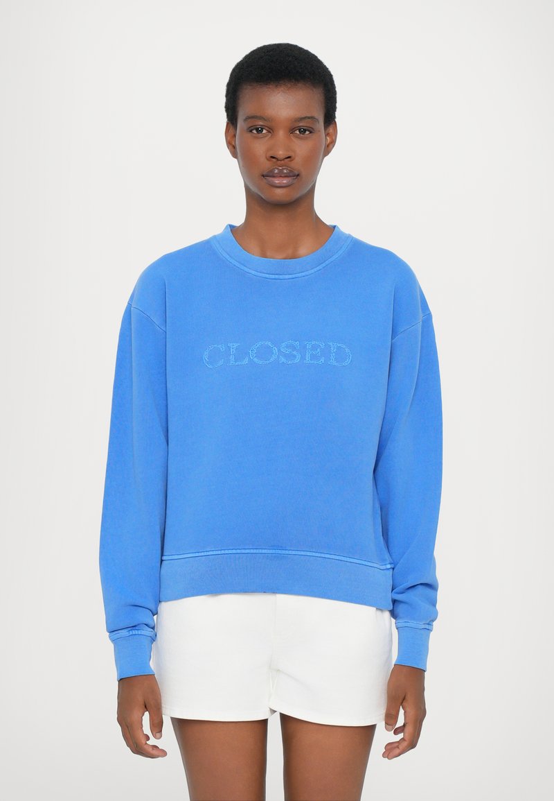 Μπλε φούτερ με το κείμενο "CLOSED" κεντημένο σε ανοιχτό χρώμα, με ριμπ μανσέτες και τελείωμα, φαρδιά εφαρμογή, φορεμένο πάνω από λευκές βερμούδες. Λείος βαμβακερός υφασμάτινος ιστός.