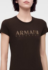 Γυναίκα φοράει εφαρμοστή μαύρη μπλούζα Armani Exchange με χρυσογράμματο λογότυπο στο στήθος.