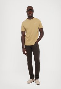 Tricou din bumbac bej cu un mic logo alb, asociat cu blugi slim-fit maro închis și mocasini bej. O șapcă maro completează ținuta.