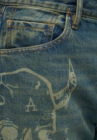 Τσέπη από τζιν με ξεθωριασμένα κίτρινα ράμματα και μερικό ανοιχτόγραφο γραφικό που μοιάζει με κεφάλι ταύρου.