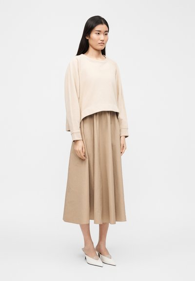 Γυναίκα με μακριά σκούρα μαλλιά, φορούσα φαρδιά μπεζ πουλόβερ, ταμπά μίντι φούστα και λευκά παπούτσια με μυτερή μύτη, στέκεται μπροστά σε απλό ανοιχτό φόντο.