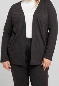 Garnitury w kolorze węgla z teksturowanym wzorem, zapinany na przód, z długimi rękawami i bocznymi kieszeniami, w zestawieniu z białym golfem z prążkami.