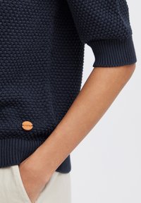 Marineblauer Strickpullover mit strukturiertem Muster, gerippten Bündchen und einem kleinen Holzlogo-Akzent. Gestylt mit Händen in den Taschen.